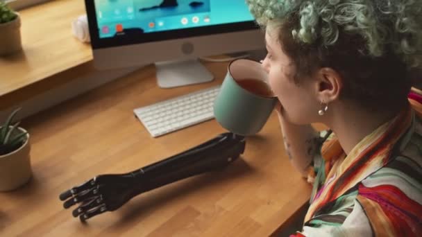 Fentről nézve, ahogy a fogyatékos kaukázusi lány teát iszik bögréből, és az ablakot nézi, ahogy a fa asztalnál ül, rajta a karprotézisével. - Felvétel, videó