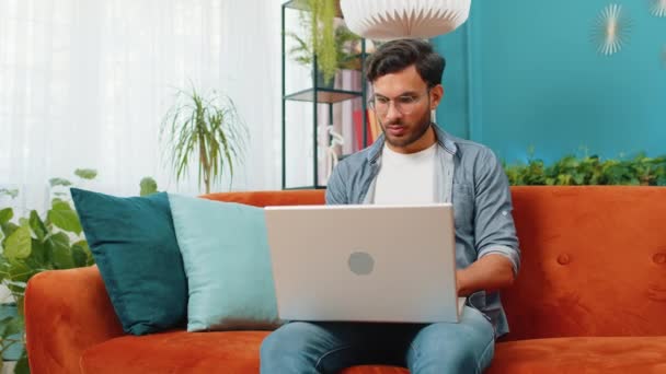 Indián férfi ül a kanapén, laptopot zárva, miután befejezte a munkát a nappaliban. Fiatal arab srác szabadúszó dolgozik online távoli munkát otthon kanapé. Elektronikus tanulás, internetes böngészés notebook számítógépen - Felvétel, videó