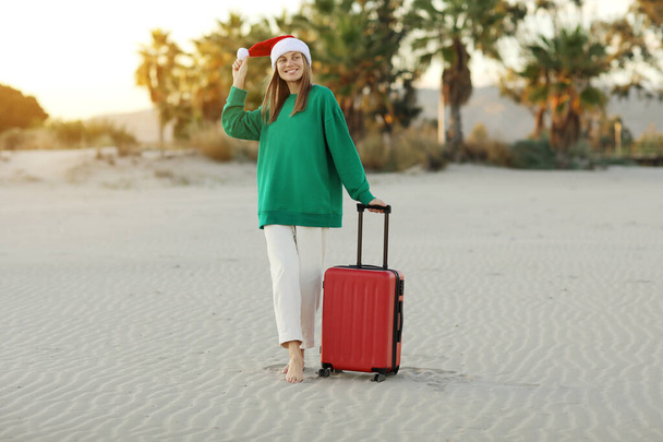 Šťastná mladá žena v klobouku Santa Claus a zelené mikiny stojí s červeným kufříkem na pláži s palmami. Vánoce a novoroční prázdniny. Oslava zimních prázdnin v zahraničí v tropech.. - Fotografie, Obrázek