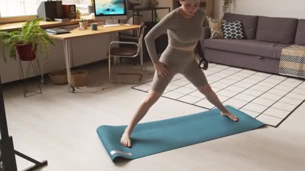 Colpo a figura intera della ragazza caucasica in forma con braccio protesico che fa stretching delle gambe sul tappeto mentre si allena a casa al mattino - Filmati, video