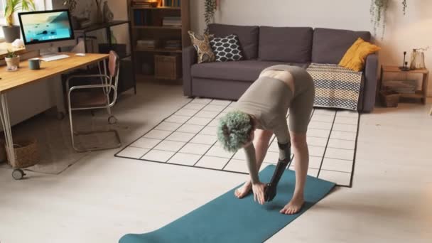 Full length shot of young Caucasian woman with arm prosthesis doing warmup exercises bend and upper twist on mat in living room during home training - Footage, Video