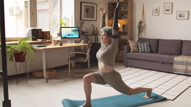 Ganzkörperaufnahme eines flexiblen kaukasischen Mädchens mit künstlichem Arm, das Yoga praktiziert, während es im gemütlichen Wohnzimmer auf einer Matte steht - Filmmaterial, Video