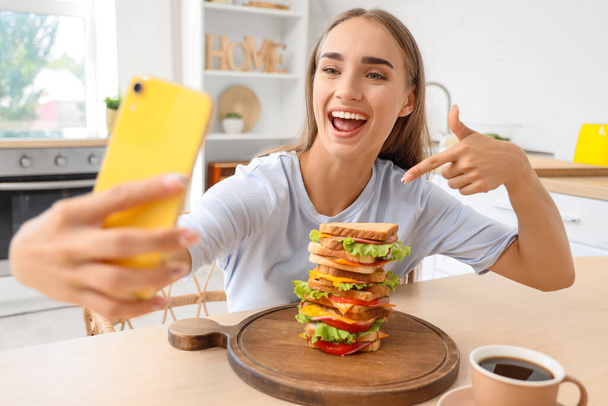 Hermosa joven tomando selfie con sabroso sándwich en la mesa en la cocina - Foto, Imagen