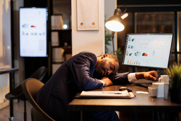 Müder leitender Angestellter, der auf dem Schreibtisch im Startup-Büro schläft, workaholischer Angestellter, der wegen Überstunden einschläft. Ausgelaugter afrikanisch-amerikanischer Geschäftsmann mit Burnout-Syndrom bei der Arbeit. - Foto, Bild