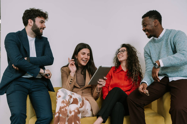 Крупним планом чотири бізнес-колеги розмовляють і посміхаються, сидячи на жовтому дивані на робочому місці - Фото, зображення