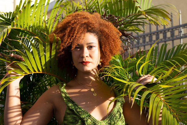 Eine Frau mit roten Haaren zwischen den Blättern einer Pflanze mit der Sonne im Gesicht am späten Nachmittag. Selbstbewusster und glücklicher Mensch. - Foto, Bild