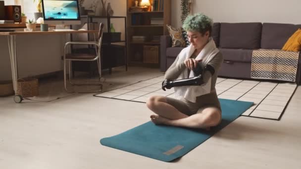 Ragazza caucasica con braccio protesico seduta sul tappetino in soggiorno a bere acqua mentre riposa dopo l'allenamento a casa - Filmati, video