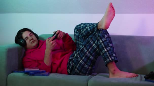 νεαρός άνδρας με ακουστικά πολύχρωμο δωμάτιο στον καναπέ που κοιμάται παίζοντας βιντεοπαιχνίδι. Υψηλής ποιότητας 4k πλάνα - Πλάνα, βίντεο