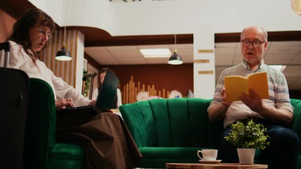 Starsza para spędzająca czas w hotelu, żona tworząca trasę podróży na laptopie, podczas gdy mąż czyta książkę w salonie. Emeryci podróżujący na międzynarodowe wakacje, czekając na odprawę. - Materiał filmowy, wideo