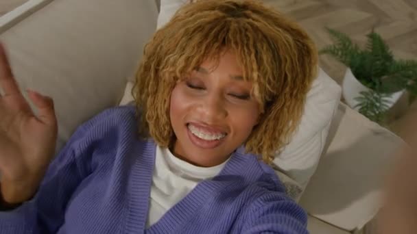 Web cam view sorridente felice afro-americano ragazza etnica donna che agita mano saluto parlare online storia di social media streaming chat sdraiato a casa divano influencer femminile cellulare videochiamata - Filmati, video