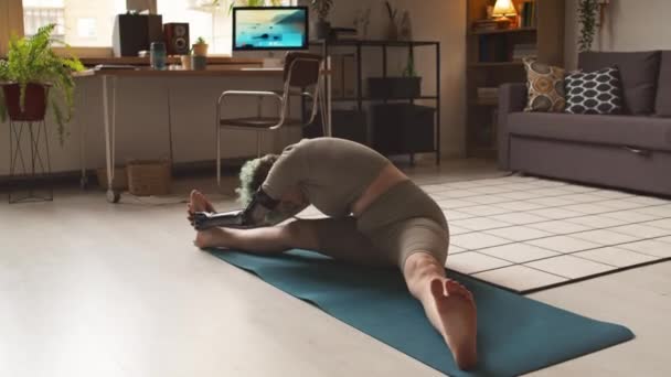 Flexibel blank meisje met armprothese zittend op mat en strekkend tot aan haar voeten, met indoor fitness training in gezellig studio appartement met warm licht - Video