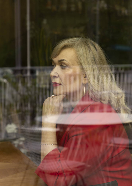 Muotokuva kaunis blondi kypsä seksuaalinen nainen istuu ikkunan takana lasi ravintola tai kahvila. Tyylikäs moderni vanhempi nainen 60-vuotiaana ajattelemassa jotain. Eläkkeelle jääminen. Pystytaso. - Valokuva, kuva