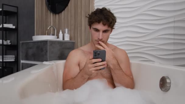 Caucasico pensoso calmo tranquillo uomo nudo rilassato ragazzo maschio seduto rilassarsi nella vasca da bagno in schiuma saponata bagno d'acqua calda premuroso cercando cellulare utilizzando smartphone pensare lettura app di social media - Filmati, video