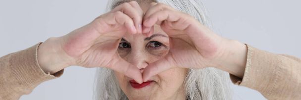 Coqueteando mujer de pelo gris mira a través del símbolo del corazón hecho de dedos sobre fondo claro. La señora madura envía amor a todos. Relación romántica - Foto, Imagen