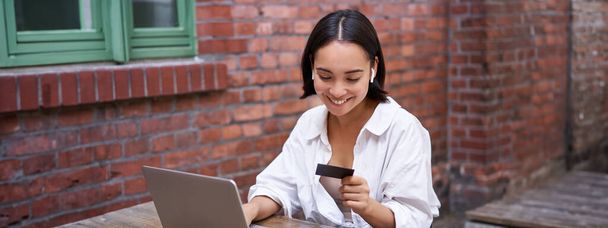 Χαμογελαστή Ασιάτισσα με φορητό υπολογιστή και ασύρματα ακουστικά, που πληρώνει με πιστωτική κάρτα, που αγοράζει online, που κάθεται με ένα φλιτζάνι καφέ. - Φωτογραφία, εικόνα