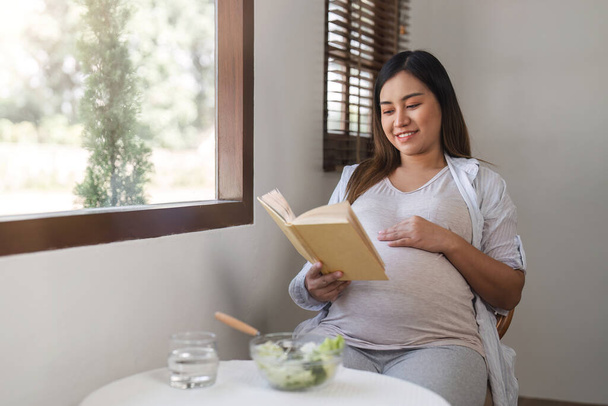 Una donna incinta felice si siede, si rilassa, legge un libro e mangia un'insalata sana sul tavolo della sala da pranzo in soggiorno... - Foto, immagini