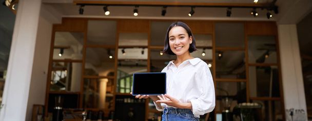 Sorrindo asiático mulher mostrando digital tablet tela, cafe proprietário mostrando smth, de pé na frente de café entrada. - Foto, Imagem