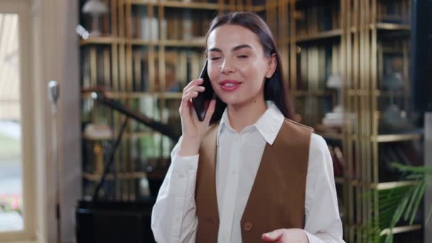 Smiling businesswoman with long hair converses on mobile phone in hotel lounge. Joyful woman discussing work via smartphone while enjoying the ambiance of the hotel lobby during her business travel. - Footage, Video