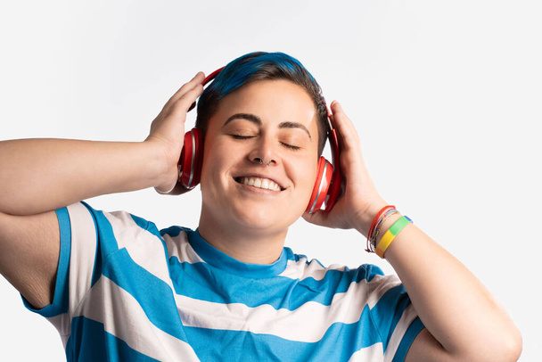 Retrato de una persona joven y andrógina con el pelo teñido de azul vibrante, con auriculares y una camisa a rayas. Están disfrutando de la música, mostrando un estilo moderno y casual con piercing en la nariz y pulseras de colores. - Foto, imagen
