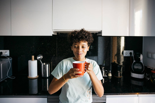 Uśmiechnięta dziesięciolatka pijąca mleko z czerwonego kubka w kuchni. Dziewczyna w domu pije kubek mleka.. - Zdjęcie, obraz