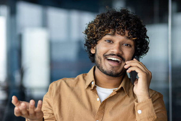 Κοντινό πορτρέτο του νεαρού επιχειρηματία, άντρας που μιλάει χαρούμενα και χαμογελαστά φιλικά και κοιτάζει την κάμερα, Ισπανόφωνος στο χώρο εργασίας μέσα στο γραφείο. - Φωτογραφία, εικόνα
