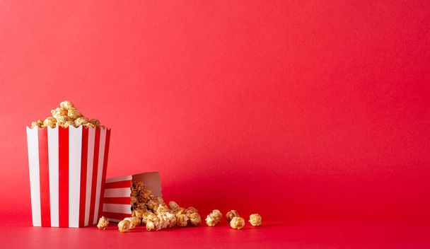 Host movie premieres with your buddies and delicious snacks. Side view photograph of tabletop featuring mouthwatering popcorn in striped boxes against red wall, providing space for movie advertisement - Photo, Image