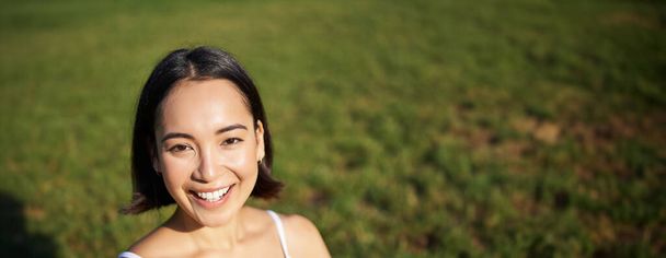 tiro vertical de jovem asiático mulher fazendo ioga, praticar mindfulness, sorrindo e olhando relaxado, sentado no tapete de borracha no parque. - Foto, Imagem