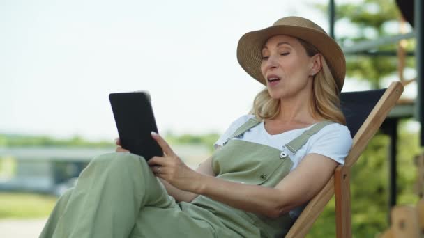 Очаровательная женщина средних лет проводит летние каникулы в сельской местности, наслаждается на открытом воздухе и расслабляется после работы в саду, сидя в кресле. Счастливая дама использует через видео-звонок в планшете - Кадры, видео