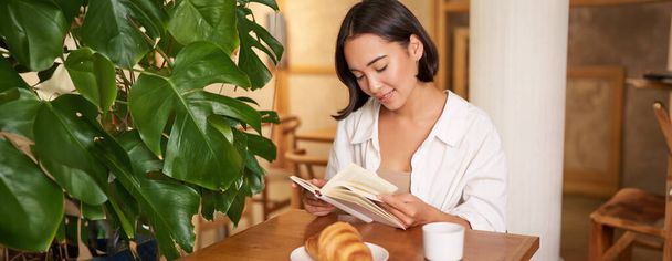 Улыбающаяся молодая азиатка наслаждается чтением книги, сидя одна в кафе, пьет кофе, смотрит на страницу. - Фото, изображение