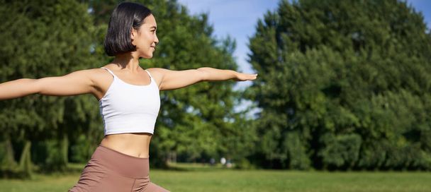 Młoda kobieta rozciągająca swoje ciało, ćwicząca i uprawiająca jogę w parku, stojąca na gumowej macie w odzieży fitness podczas sesji treningowej. - Zdjęcie, obraz