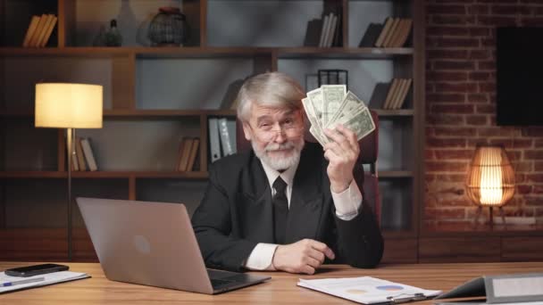 Pozytywny starszy mężczyzna w formalnym garniturze i okulary wskazujące na fankę papierowych pieniędzy podczas pobytu w wygodnym biurze. Profesjonalne biznesowe mrugnięcie okiem przed kamerą, chwaląc się zyskiem. - Materiał filmowy, wideo