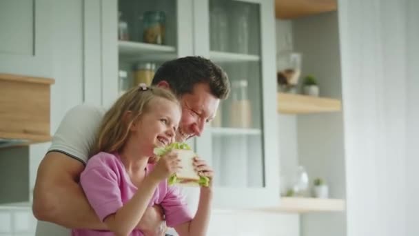 Heureux père riant tient et balance fille avec sandwich dans les mains, se tient dans la cuisine. Une atmosphère de joie et de positivité dans la maison. Le concept de l'enfance heureuse et de la parentalité. Haute qualité - Séquence, vidéo