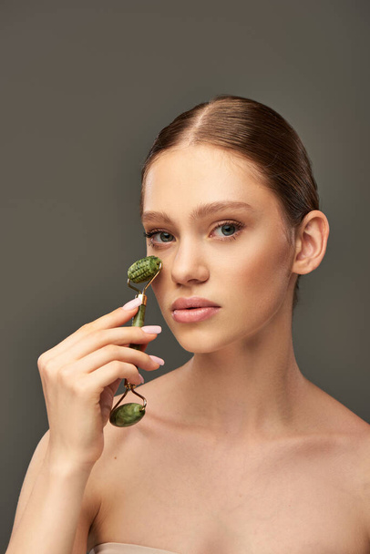 young woman with perfect skin massaging face with jade roller on grey background, skin rejuvenation - Photo, Image