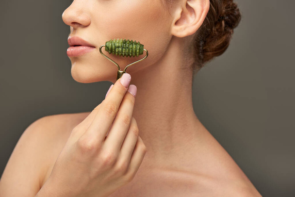 bijgesneden vrouw met perfecte huid masseren gezicht met jade roller op grijze achtergrond, huidverzorging - Foto, afbeelding