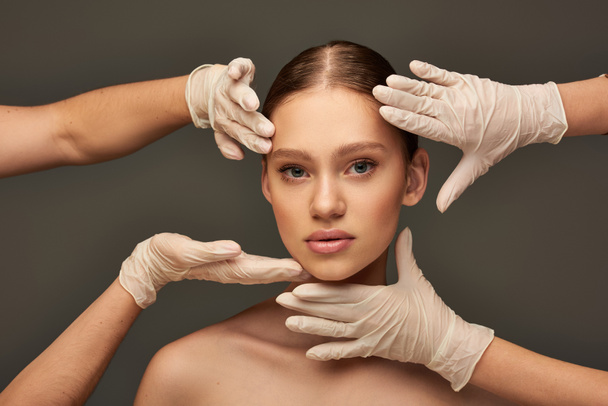 Schönheitschirurgen in medizinischen Handschuhen untersuchen das Gesicht einer jungen Frau auf grauem Hintergrund, Gesichtsbehandlung - Foto, Bild