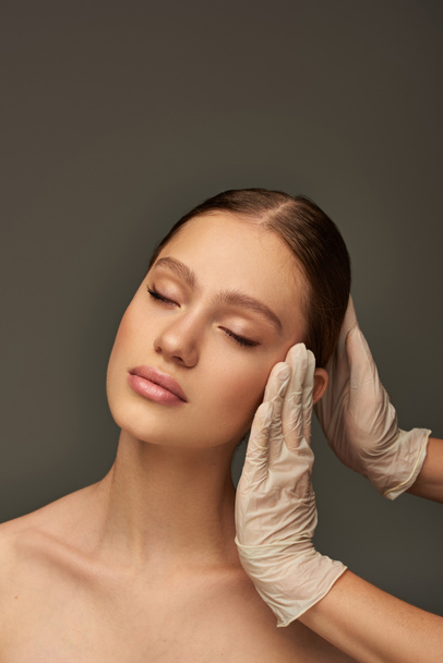 esteticista en guantes médicos tocando la cara de mujer joven sobre fondo gris, tratamiento facial - Foto, imagen
