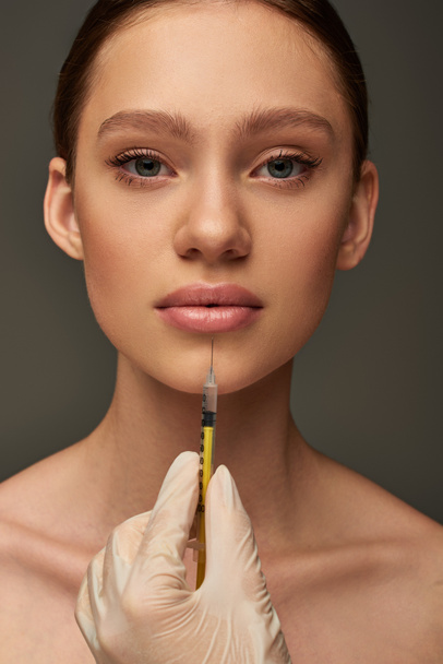 esthetician in medical glove holding syringe near young woman on grey background, lip filler - Photo, Image