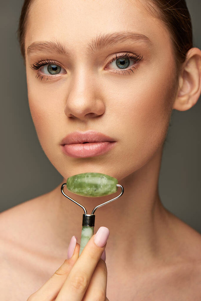 young woman doing face massage with green jade roller on grey background, skin rejuvenation - Photo, Image