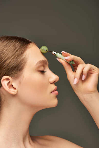 side view of young woman with closed eyes doing face massage with jade roller on grey background - Photo, Image