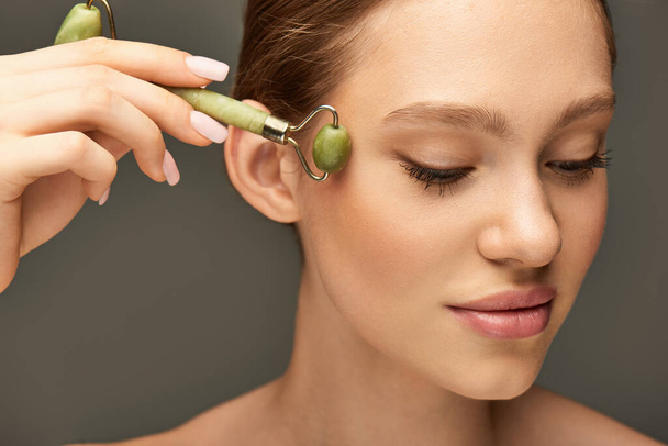 portrait of young woman doing face massage with green jade roller on grey background, self-care - Photo, Image