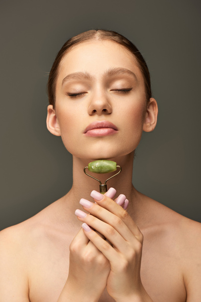 portrait of young woman doing massage of neck with green jade roller on grey background, self-care - Photo, Image