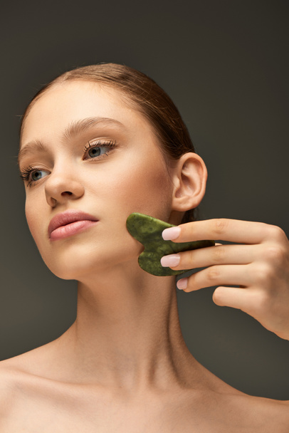 jovem fazendo massagem facial com jade gua sha stone no fundo cinza, conceito de contorno facial - Foto, Imagem