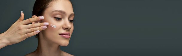 beautiful young woman with bare shoulders applying cream on face on grey background, banner - Photo, Image