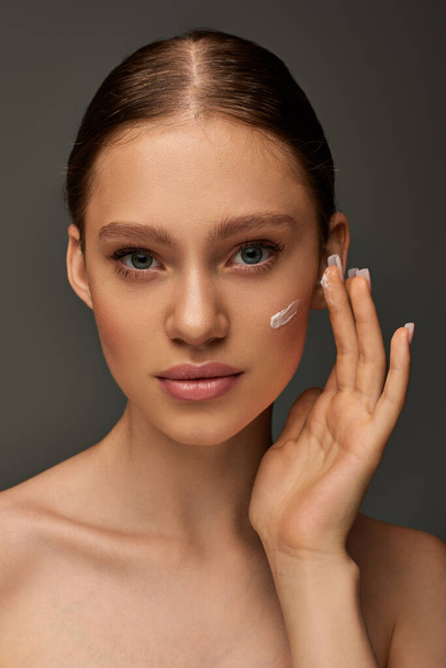 beautiful young woman with bare shoulders applying cream on face on grey background, look at camera - Fotografie, Obrázek