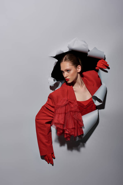 bonita mujer joven en chaqueta roja y guantes rompiendo a través del agujero en el fondo gris, conceptual - Foto, imagen