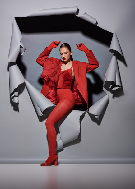 powerful young woman in red outfit with gloves breaking through torn grey background with hole - Photo, Image