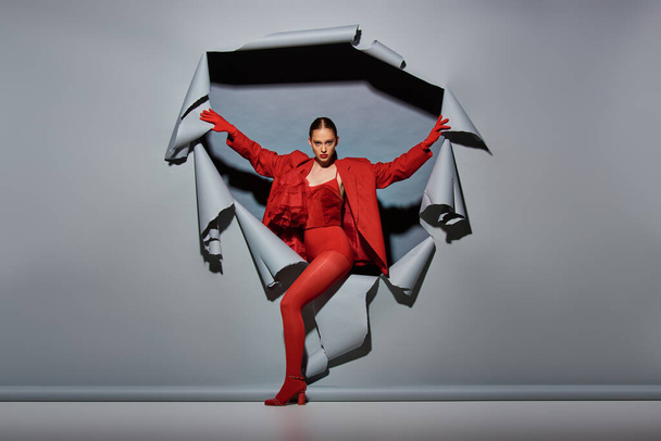 mujer joven con confianza en traje rojo con guantes rompiendo a través de fondo gris roto con agujero - Foto, imagen