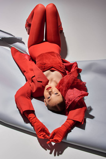 full length of young model in red outfit with tights posing lying on grey background, look at camera - Photo, Image