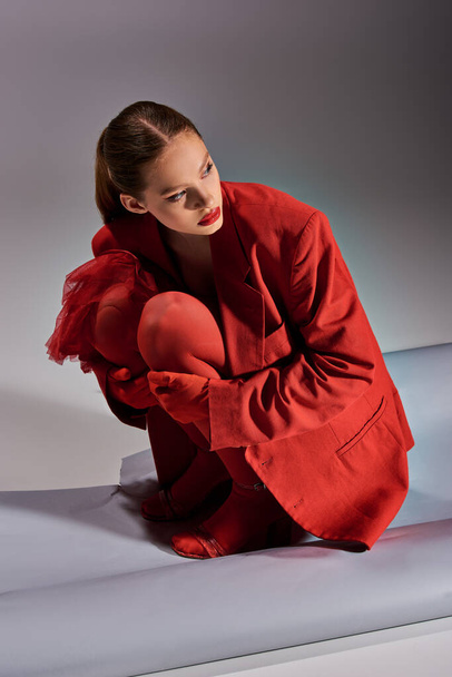 stylowy model w czerwonym stroju z rajstopami i szpilkami siedzącymi i odwracającymi wzrok na szarym tle - Zdjęcie, obraz