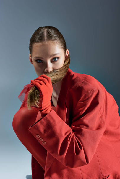 stylová mladá žena v červeném oblečení s rukavicemi tahání culík na šedém pozadí, podívejte se na kameru - Fotografie, Obrázek